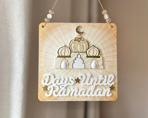 Gold white ivory plaque with mini mosque adorned with beads. Writing that says: Days until Ramadan, and it includes a few gold stars. Gold twine to hang, with beads on either side.