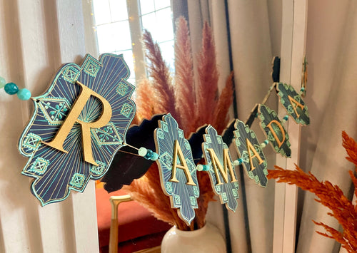 ramadan banner made of wood material and beads. each flag has a letter with blue, gold and teal and mint green hand painted details. they flags are strung together with gold twine and are hung on a mirror in this image.