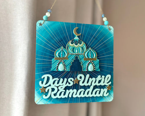 Teal mint green plaque with mini mosque adorned with beads. Writing that says: Days until Ramadan, and it includes a few gold stars. Gold twine to hang, with beads on either side.