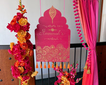 Load image into Gallery viewer, A festive Mehndi backdrop setup featuring a bright pink arch banner with gold ornamental detailing, draped pink fabric, a tall floral garland in red, orange, and yellow, a matching floral arrangement in a vase, and draped orange fabric on the floor.