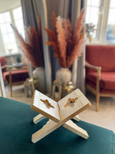 Load image into Gallery viewer, Open book on a stand in a living room setting with decor elements.
