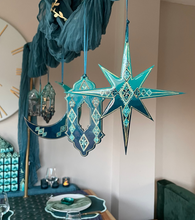 Load image into Gallery viewer, Decorative hanging stars and crescent moons on a wall with a table setting in the foreground.