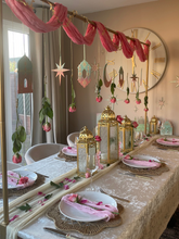 Load image into Gallery viewer, Decorative table setting with lanterns and hanging ornaments in a room with a clock on the wall.