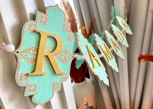 ramadan banner made of wood material and beads.  each flag has a letter with gold and pink and ivory hand painted details. they flags are strung together with gold twine and are hung on a mirror in this image.