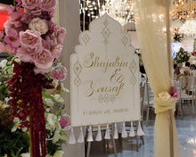 Load image into Gallery viewer, A wedding walima nikka reception venue entrance with a decorative white arch sign  in gold script, draped with cream fabric, beside a large pink and burgundy floral arrangement on a gold stand.