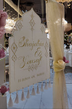 Load image into Gallery viewer, A decorative wedding sign hanging from a gold frame, with cream draped fabric and tassels, in a decorated event venue. 
