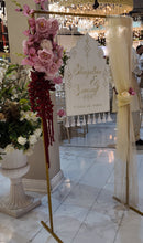 Load image into Gallery viewer, A wedding reception venue with a decorative entrance sign on a gold stand, next to a cream draped fabric stand. The background shows a man in traditional South Asian attire standing in a decorated hall with white marble floors, white tablecloths, and hanging crystal chandeliers.